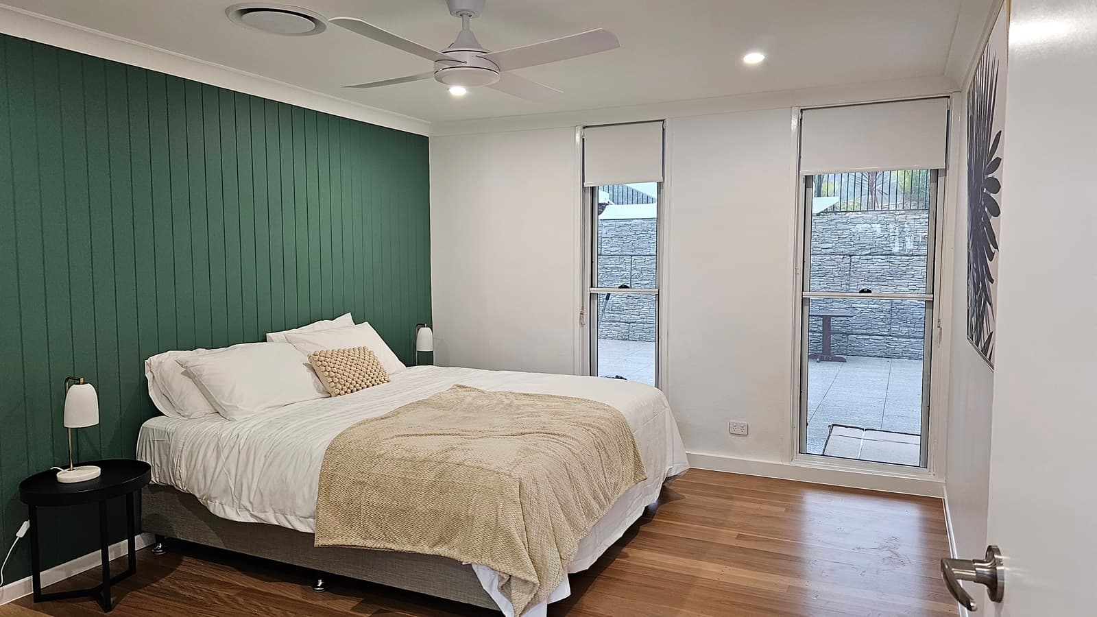 On the left, there is a green panelled wall with a double bed against it, featuring white bedding, a beige throw, and a beige cushion. A small round black side table with a white lamp is next to the bed. On the right, two tall windows with white blinds overlook a stone wall outside. The floor is wooden, and the ceiling is white with recessed lighting, a ceiling fan, and a circular air vent.
