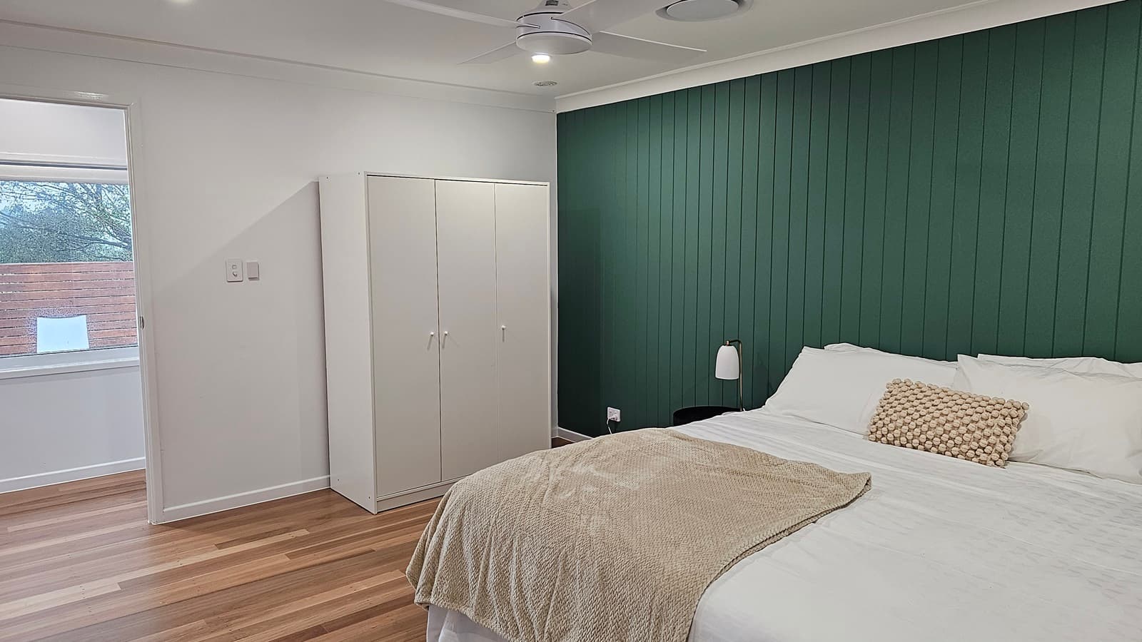 On the left, there is a white wardrobe next to a window with a white blind, showing a view of trees and a wooden fence outside. On the right, a double bed with white bedding, a beige throw, and a beige cushion is positioned against a green panelled wall. The floor is wooden with alternating light and dark planks, and the ceiling is white with a ceiling fan, recessed lighting, and circular air vents.