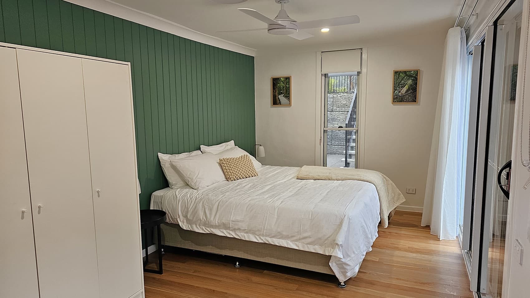 On the left, there is a white wardrobe next to a green panelled wall. In the middle, a double bed with white bedding, a beige throw, and a beige cushion. On the right, a tall window with a white blind overlooks a stone wall and stairs outside. The floor is wooden, and the ceiling is white with a ceiling fan and recessed lighting.
