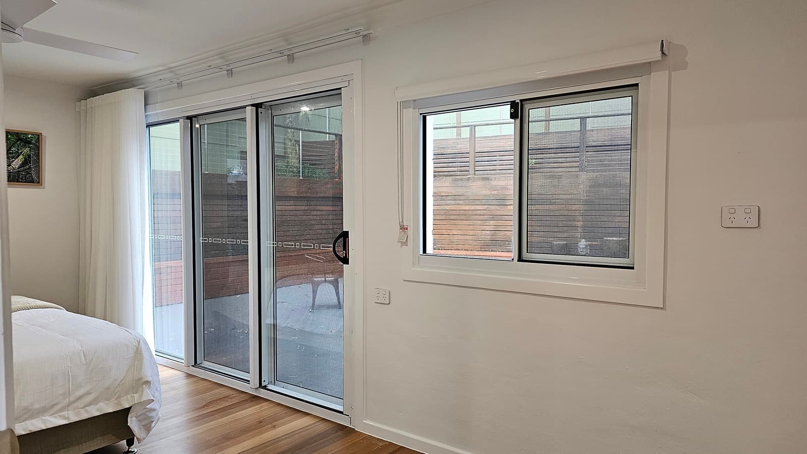 On the left, there is a bed with white bedding partially visible, next to a large sliding glass door with white curtains. On the right, a white wall with a rectangular window featuring a white blind and a view of a wooden fence outside. The floor is wooden with light and dark planks, and the ceiling is white with a recessed light and a ceiling fan.