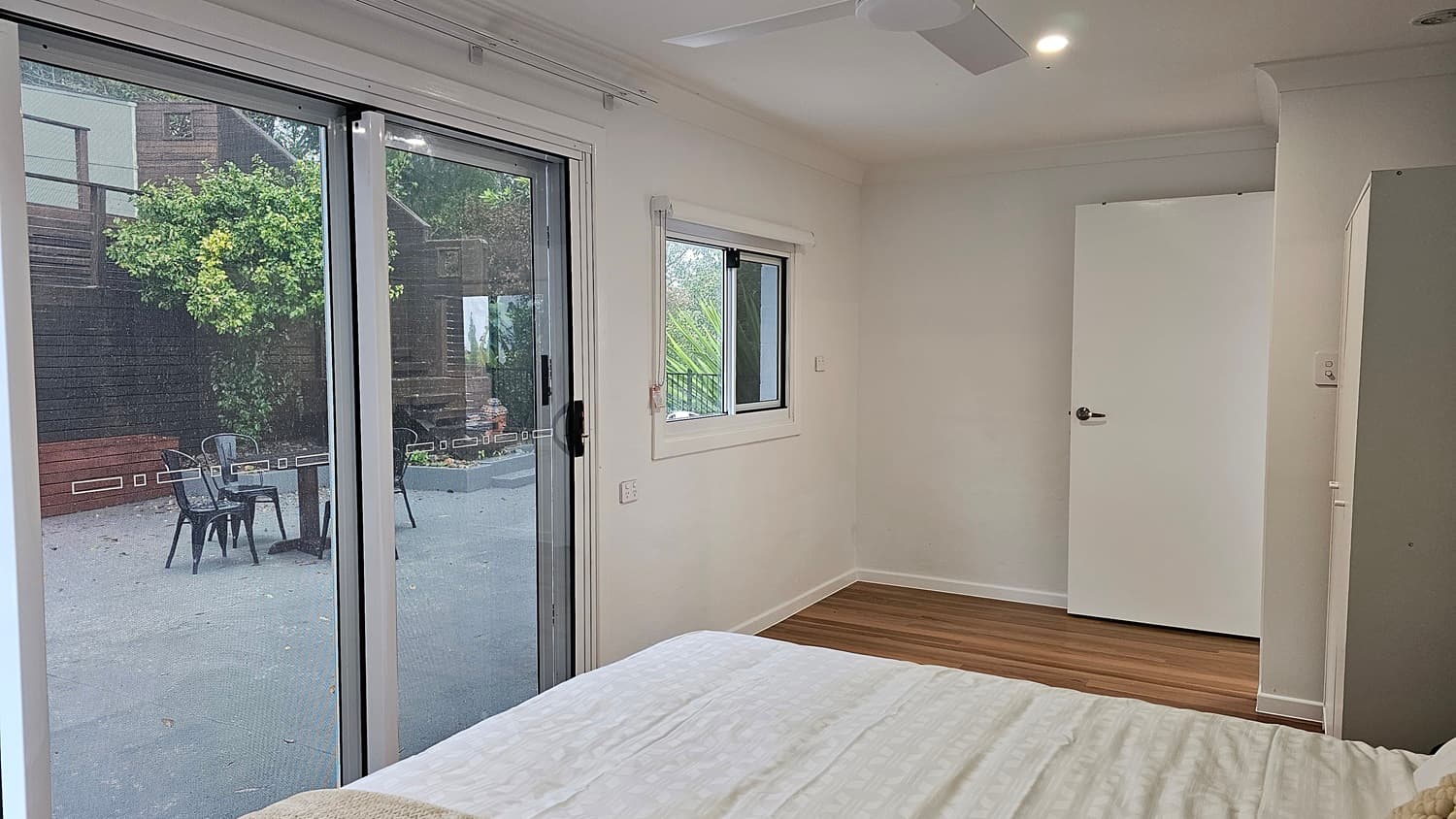 On the left, there is a large sliding glass door with white curtains, leading to an outdoor patio with black chairs and a table. In the middle, a white bed with a beige throw is partially visible. On the right, a white wall with a small rectangular window with a white blind, and a closed white door. The floor is wooden with light and dark planks, and the ceiling is white with a ceiling fan and recessed lighting.