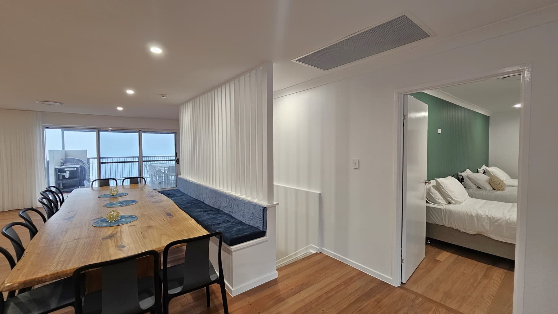On the left, there is a long wooden dining table with black chairs and blue placemats, next to large sliding glass doors leading to a balcony with outdoor furniture. In the middle, a white partition with vertical slats and a built-in blue bench. On the right, an open doorway reveals a bedroom with a green panelled wall and a bed with white bedding. The floor is wooden, and the ceiling is white with recessed lighting and an air vent.
