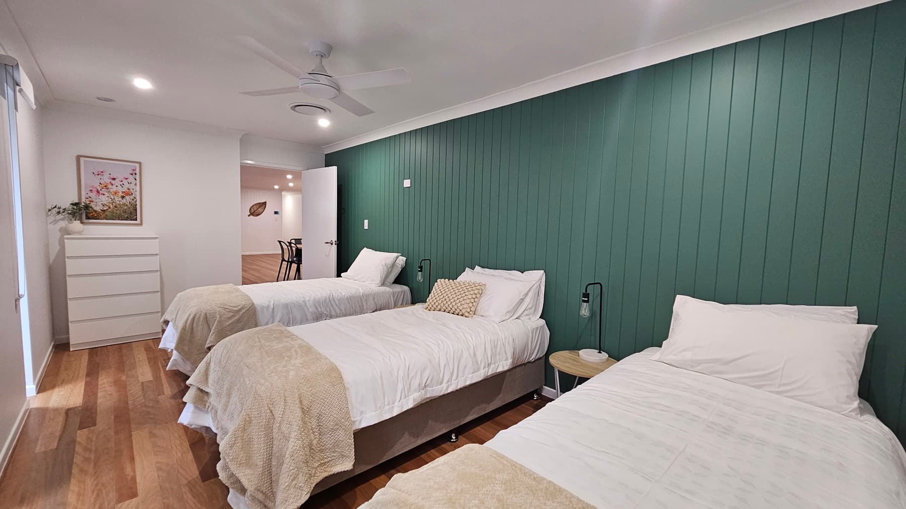 On the left, there is a white chest of drawers with a potted plant and a framed floral picture above it. In the middle, three single beds with white bedding and beige throws are aligned against a green panelled wall. On the right, a small round wooden side table with a lamp is next to the bed. The floor is wooden, and the ceiling is white with recessed lighting, a ceiling fan, and an air vent.