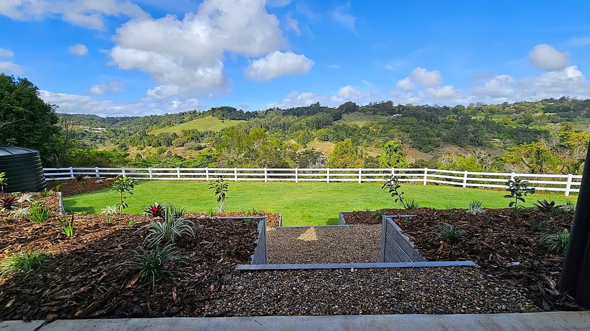 On the left, there is a white fence running along a green lawn with various plants and shrubs in a mulched garden bed. In the middle, a concrete patio extends from the house, supported by black metal posts. On the right, a sliding glass door leads to the interior of the house, and a rocky landscaped area with trees is visible. The floor is concrete, and the ceiling of the patio is white with recessed lighting.