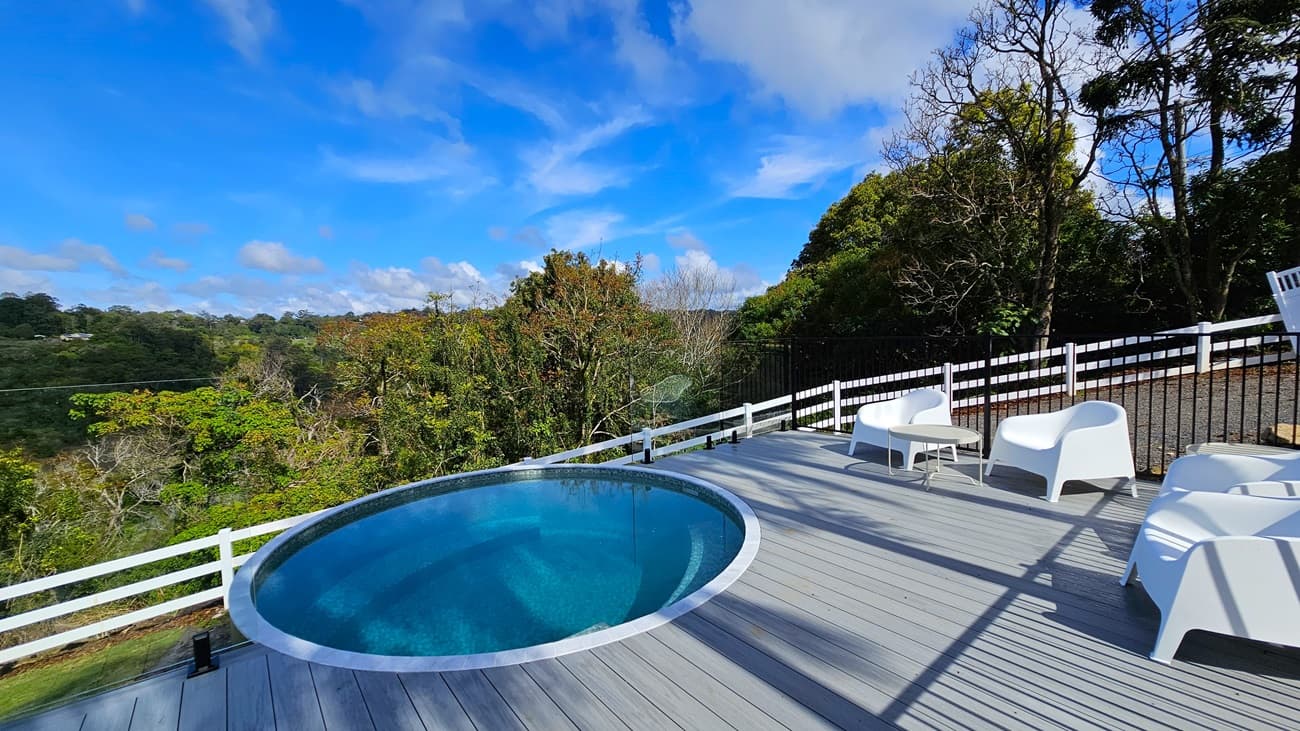 On the left, there is a round above-ground pool with clear blue water, surrounded by a white railing. In the middle, a grey wooden deck extends from the pool area. On the right, there are white plastic chairs and a small white table on the deck, with a black metal fence and white wooden fence behind them. The background features dense green trees and foliage. The floor is made of grey wooden planks, and there is no visible ceiling.