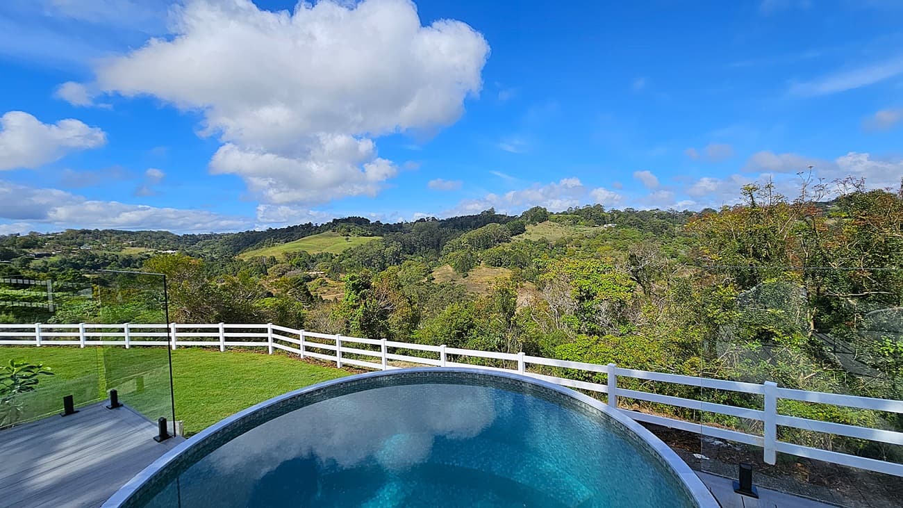 On the left, there is a glass fence panel next to a grey wooden deck. In the middle, a round above-ground pool with clear blue water is bordered by a white railing. On the right, a white wooden fence runs along the edge of a green lawn. The background features rolling green hills and dense trees. The floor is a combination of grey wooden decking and green grass.