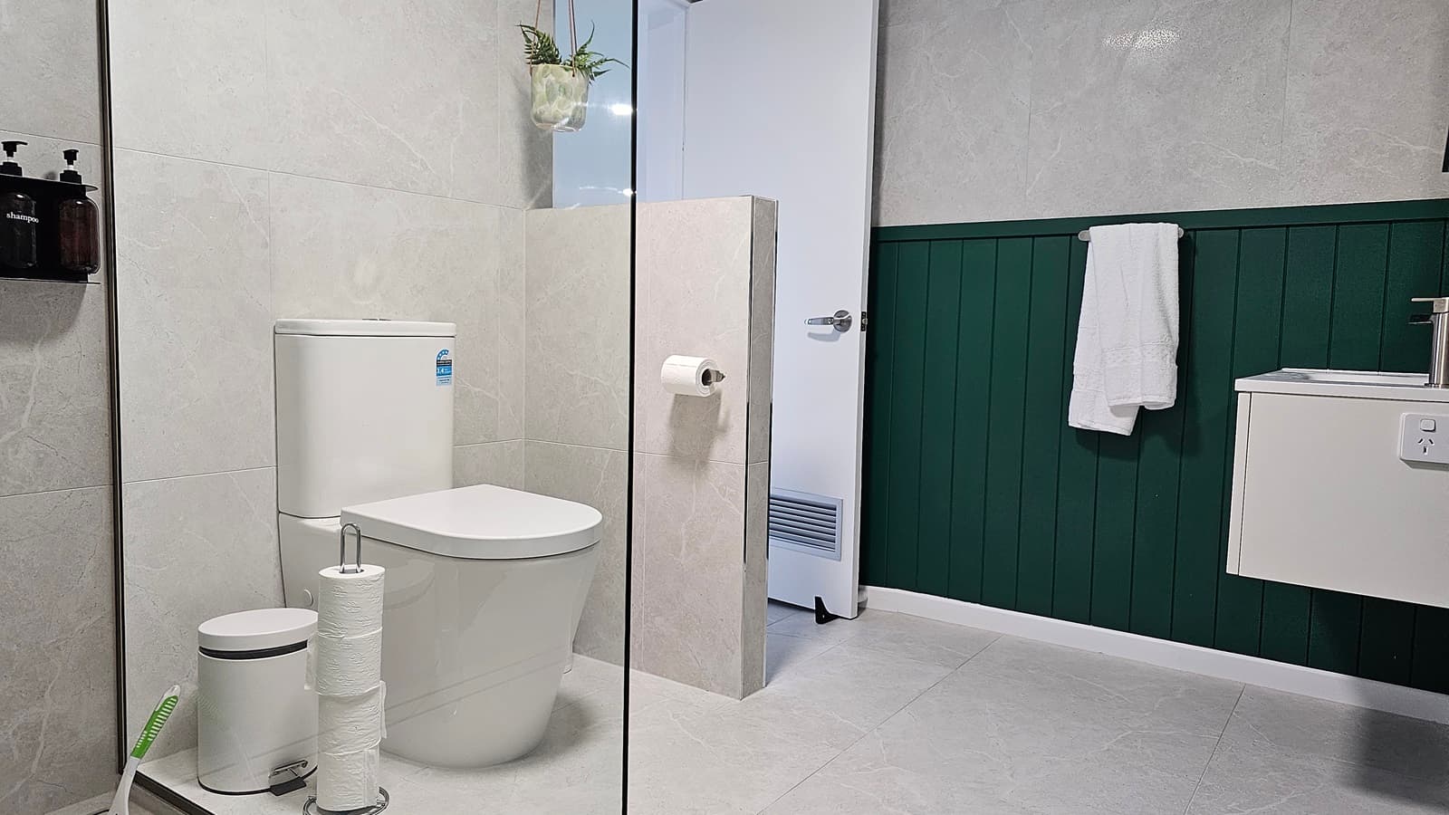 On the left, there is a white toilet with a small white bin and a toilet roll holder on the floor. In the middle, a partial wall with a mounted toilet roll holder separates the toilet area from the rest of the room. On the right, a white sink with a silver tap is mounted on a green panelled wall, with a white towel hanging on a rail. The floor is covered in light grey tiles, and the ceiling is white. A hanging plant is above the toilet, and a white door is in the background.