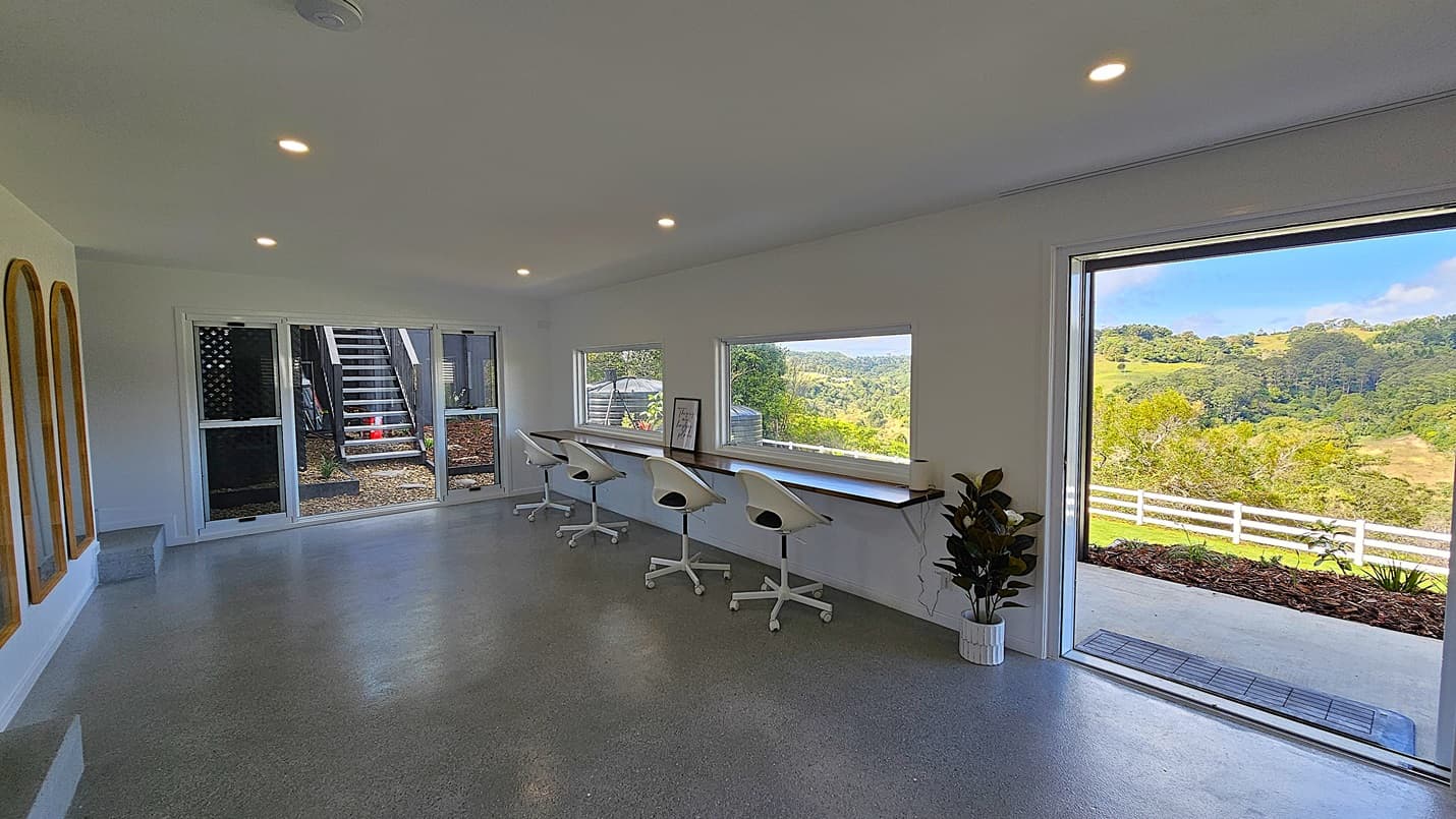 On the left, there are three arched mirrors mounted on a white wall. In the middle, a set of glass doors opens to an outdoor staircase. On the right, a long wooden desk with four white swivel chairs is positioned against a wall with two large windows, offering a view of a green landscape with hills and trees. The foreground features a polished concrete floor, and the ceiling is white with recessed lighting. A potted plant is placed near the open sliding door on the right.