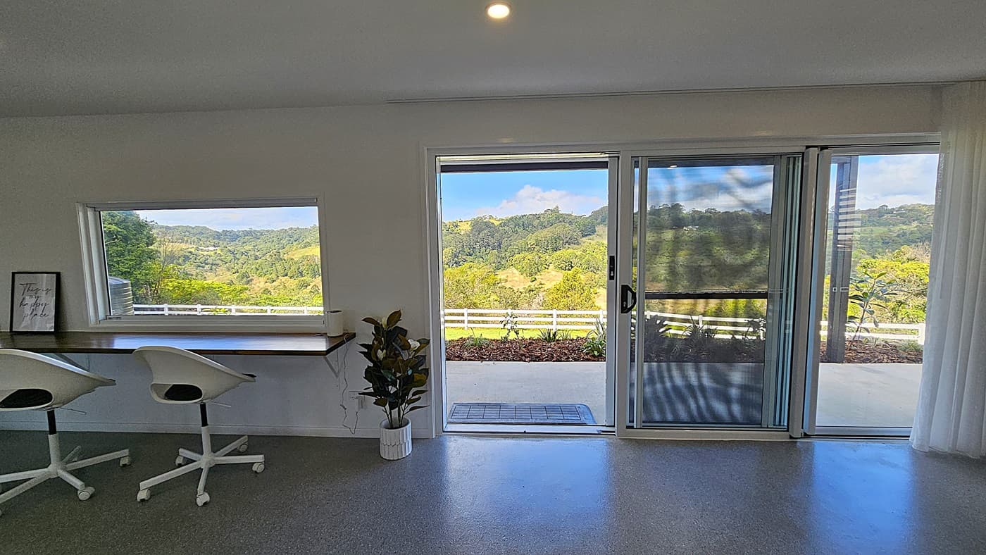 On the left, there is a long wooden desk with two white swivel chairs and a framed picture. In the middle, a large sliding glass door opens to a patio with a view of green hills and trees. On the right, another large window with white curtains offers a similar view. The foreground features a grey carpeted floor, and the ceiling is white with recessed lighting. A potted plant is placed near the sliding door.