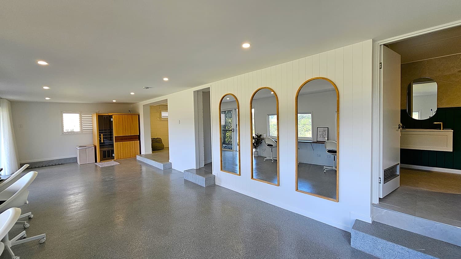 On the left, there are white swivel chairs and a small wooden sauna next to a window with white blinds. In the middle, three arched mirrors with wooden frames are mounted on a white panelled wall. On the right, an open door reveals a bathroom with a white sink and a round mirror above it. The background includes a small step leading to another room with a desk and chair. The floor is polished concrete, and the ceiling is white with recessed lighting.