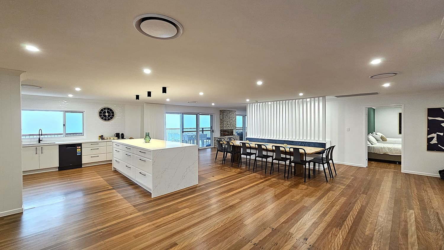On the left, there is a modern kitchen with white cabinets, a black dishwasher, a black sink, and a large clock on the wall. In the middle, a large white marble island with drawers and a few decorative items on top. On the right, a long wooden dining table with black chairs is positioned against a wall with vertical white blinds. The floor is polished wooden planks, and the ceiling is white with recessed lighting and circular air vents. The background includes a doorway leading to a bedroom.