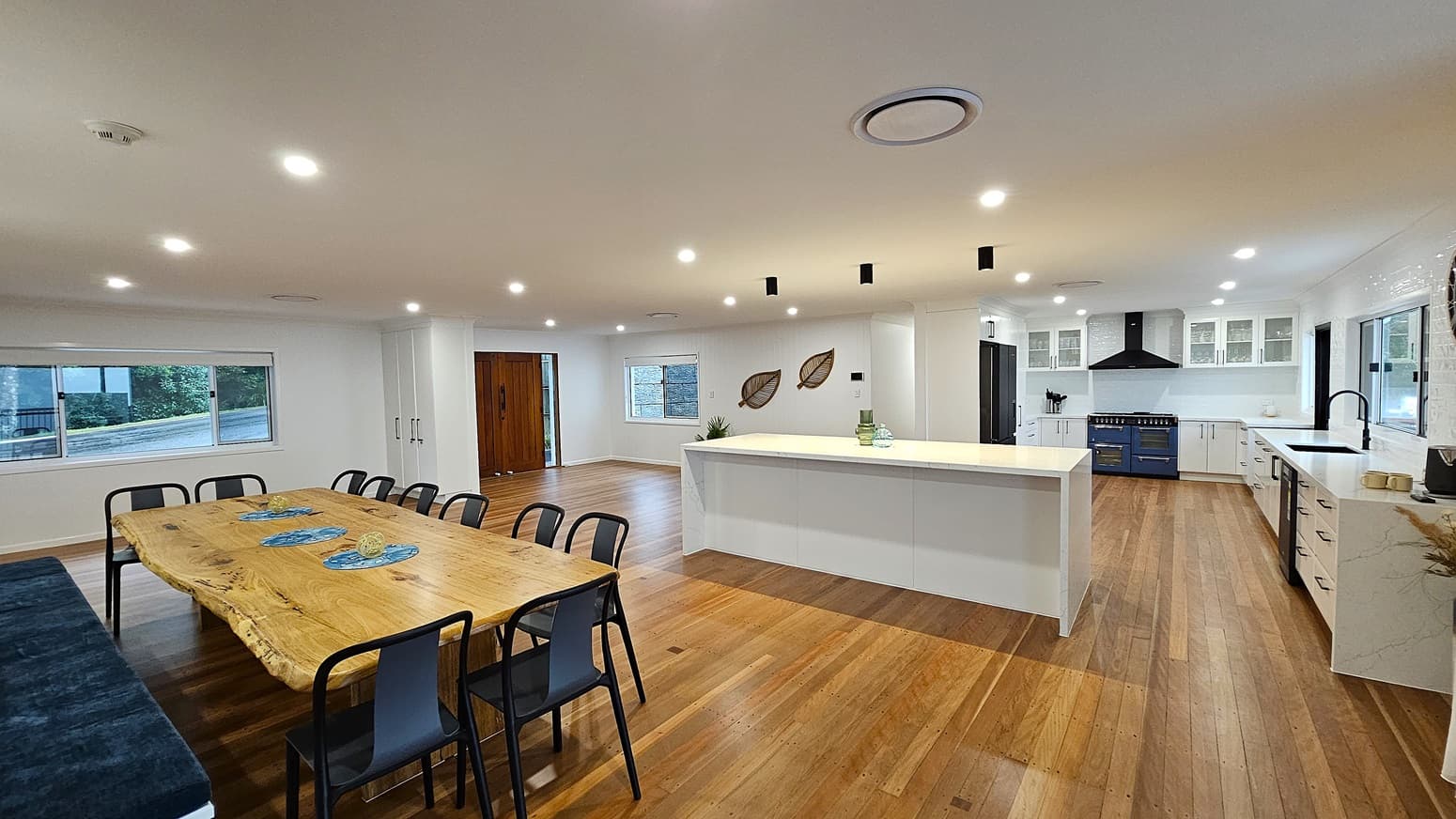 On the left, there is a long wooden dining table with black chairs, positioned near a large window with white blinds. In the middle, a spacious kitchen features a large white island countertop with decorative items. On the right, white cabinets with black handles, a black sink, and a blue oven with a black range hood. The floor is polished wooden planks, and the ceiling is white with recessed lighting and black pendant lights.