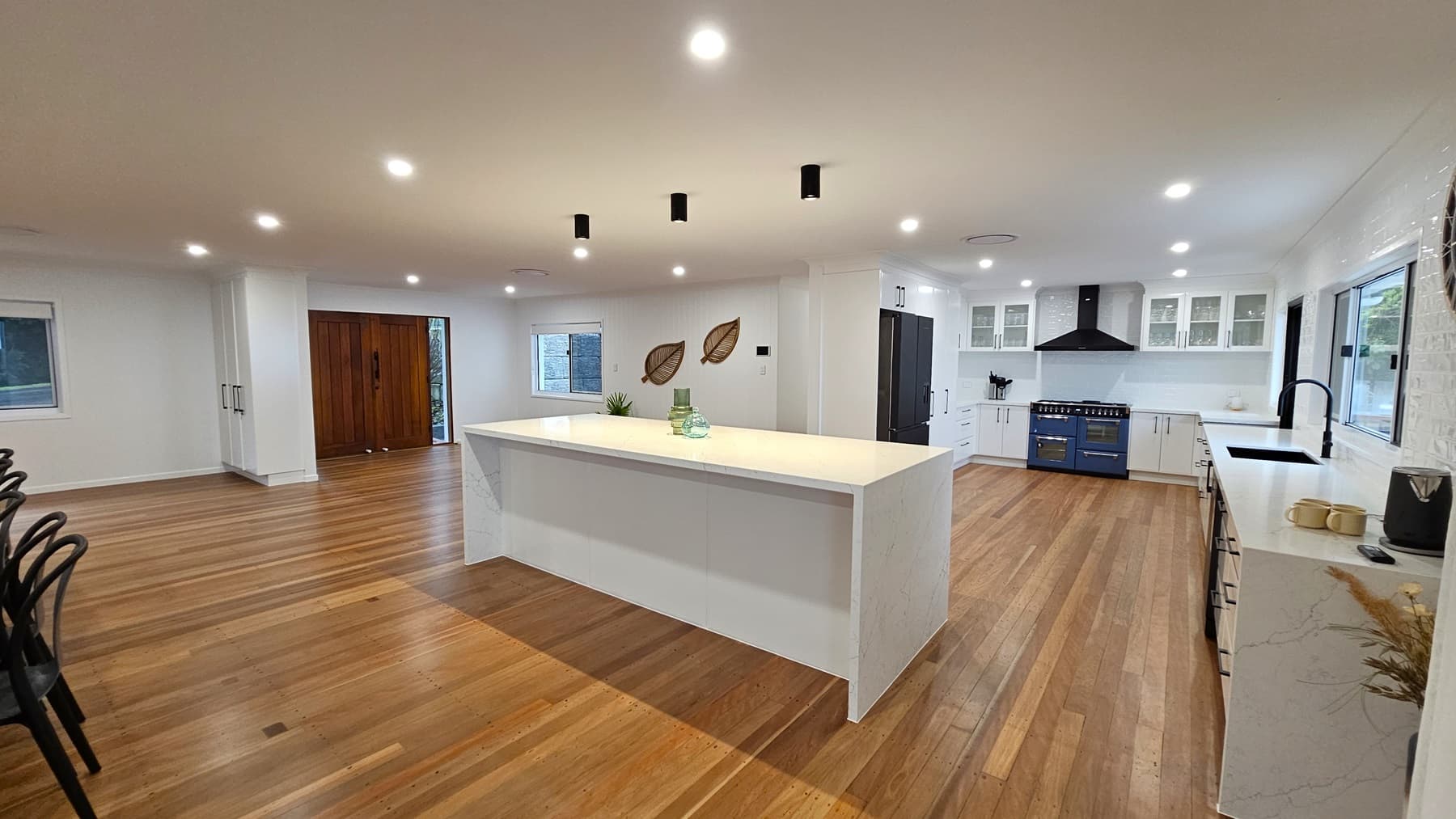 On the left, there is a long wooden dining table with black chairs near a large window with white blinds. In the middle, a spacious kitchen features a large white island countertop with decorative items. On the right, white cabinets with black handles, a black sink, and a blue oven with a black range hood. The floor is polished wooden planks, and the ceiling is white with recessed lighting and black pendant lights. The background includes large wooden doors and decorative leaf wall hangings.
