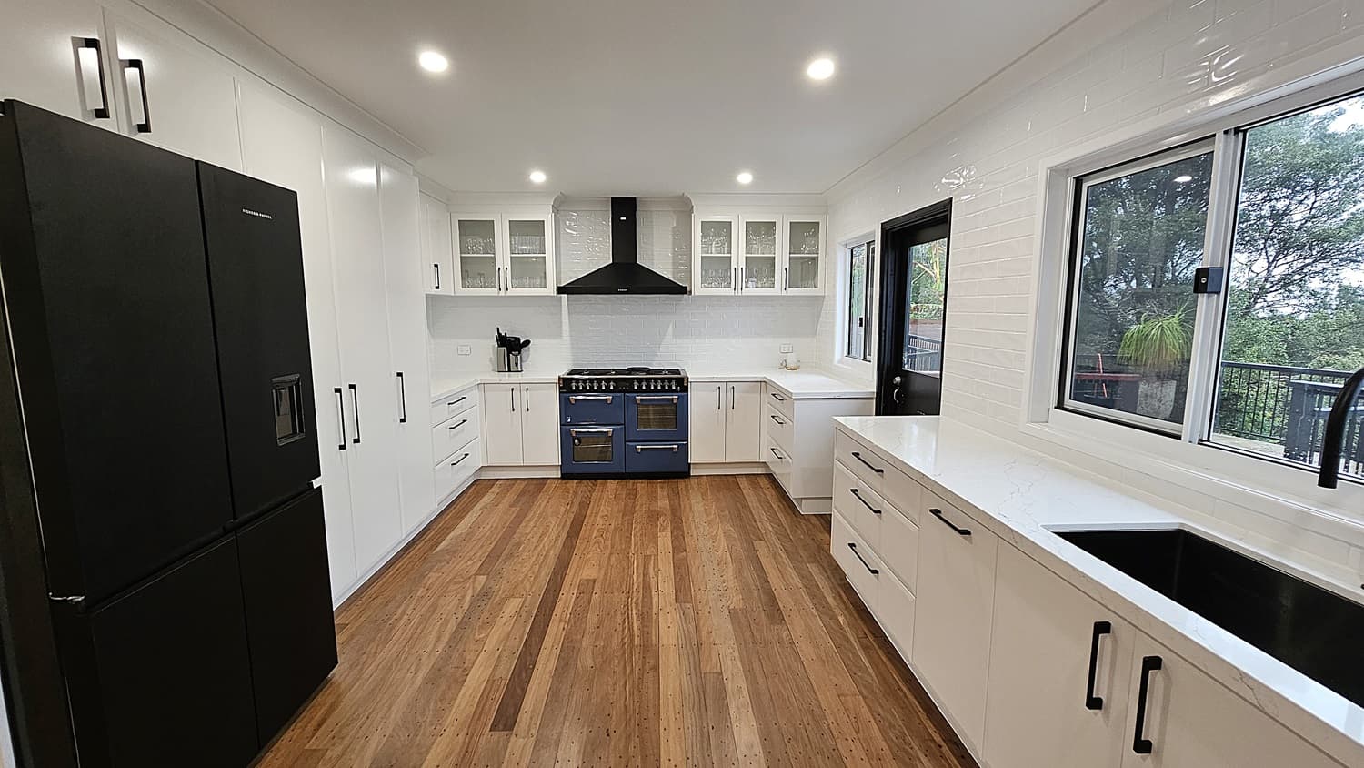 On the left, there is a large black refrigerator next to white cabinets with black handles. In the middle, a blue oven with a black range hood is flanked by white countertops and cabinets. On the right, a white countertop with black handles runs beneath a large window and a black sink. The floor is polished wooden planks, and the ceiling is white with recessed lighting. The background includes a glass door leading to an outdoor area with trees visible through the window.