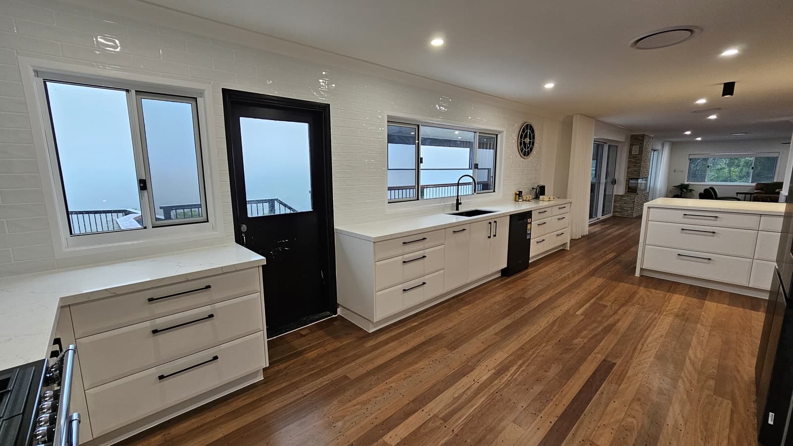 On the left, there is a white countertop with drawers beneath a window and a black door with a glass panel. In the middle, a long white countertop with a black sink and dishwasher beneath another window. On the right, a large white island with drawers and a clock on the wall above the countertop. The floor is polished wooden planks, and the ceiling is white with recessed lighting and a circular air vent. The background includes a living area with large windows and a sliding glass door.