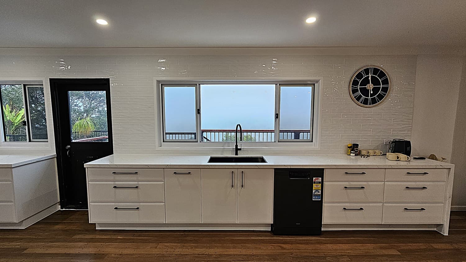 On the left, there is a black door with a glass panel next to a white countertop and a window. In the middle, a long white countertop with a black sink and a dishwasher beneath a large window. On the right, a white countertop with drawers, a black coffee maker, mugs, and a large black clock on the wall. The floor is polished wooden planks, and the ceiling is white with recessed lighting. The background includes a view of trees and a railing through the windows.