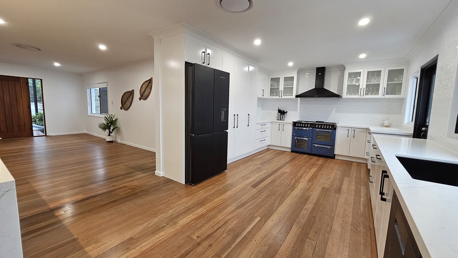 On the left, there is a wooden entrance door with glass panels and a small potted plant near a window. In the middle, a black refrigerator is surrounded by white cabinets with black handles. On the right, a blue oven with a black range hood is flanked by white countertops and cabinets, with a black sink under a window. The floor is polished wooden planks, and the ceiling is white with recessed lighting and a circular air vent. The background includes decorative leaf wall hangings.