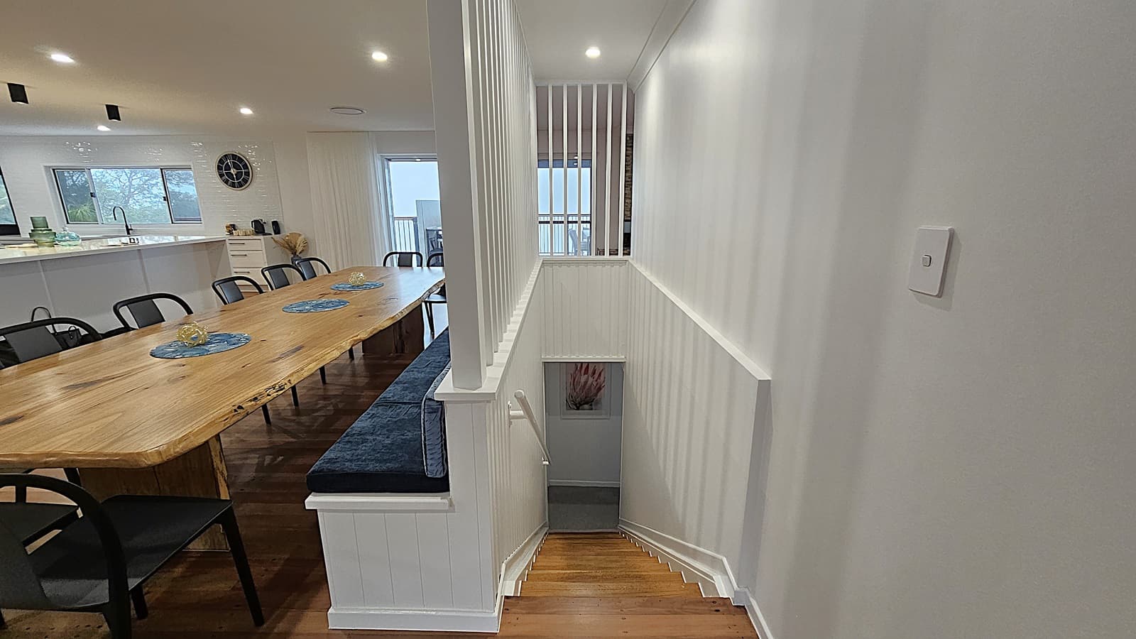 On the left, there is a long wooden dining table with black chairs and blue placemats, positioned near a white kitchen island with a black sink and decorative items. In the middle, a staircase with white railings leads down to a lower level, with a framed picture on the wall at the bottom. On the right, a white wall with a light switch. The floor is polished wooden planks, and the ceiling is white with recessed lighting.