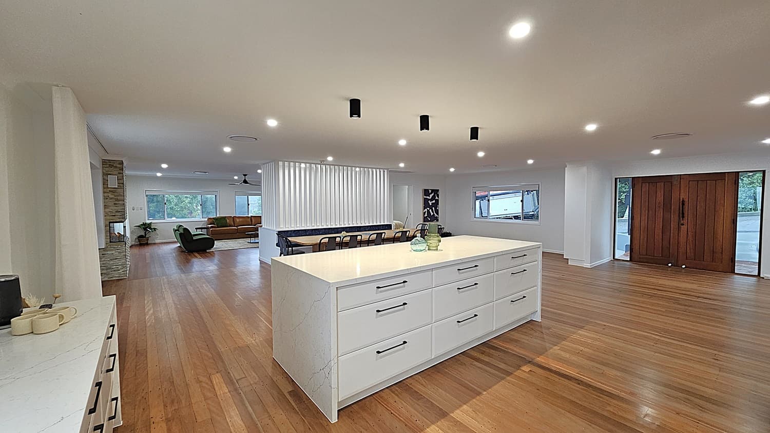 On the left, there is a white countertop with mugs and a stone fireplace next to a green armchair. In the middle, a large white kitchen island with drawers and decorative items on top. On the right, a set of wooden double doors with glass panels. The floor is polished wooden planks, and the ceiling is white with recessed lighting and black pendant lights. The background includes a dining table with black chairs, a tan sofa, and large windows with views of trees.