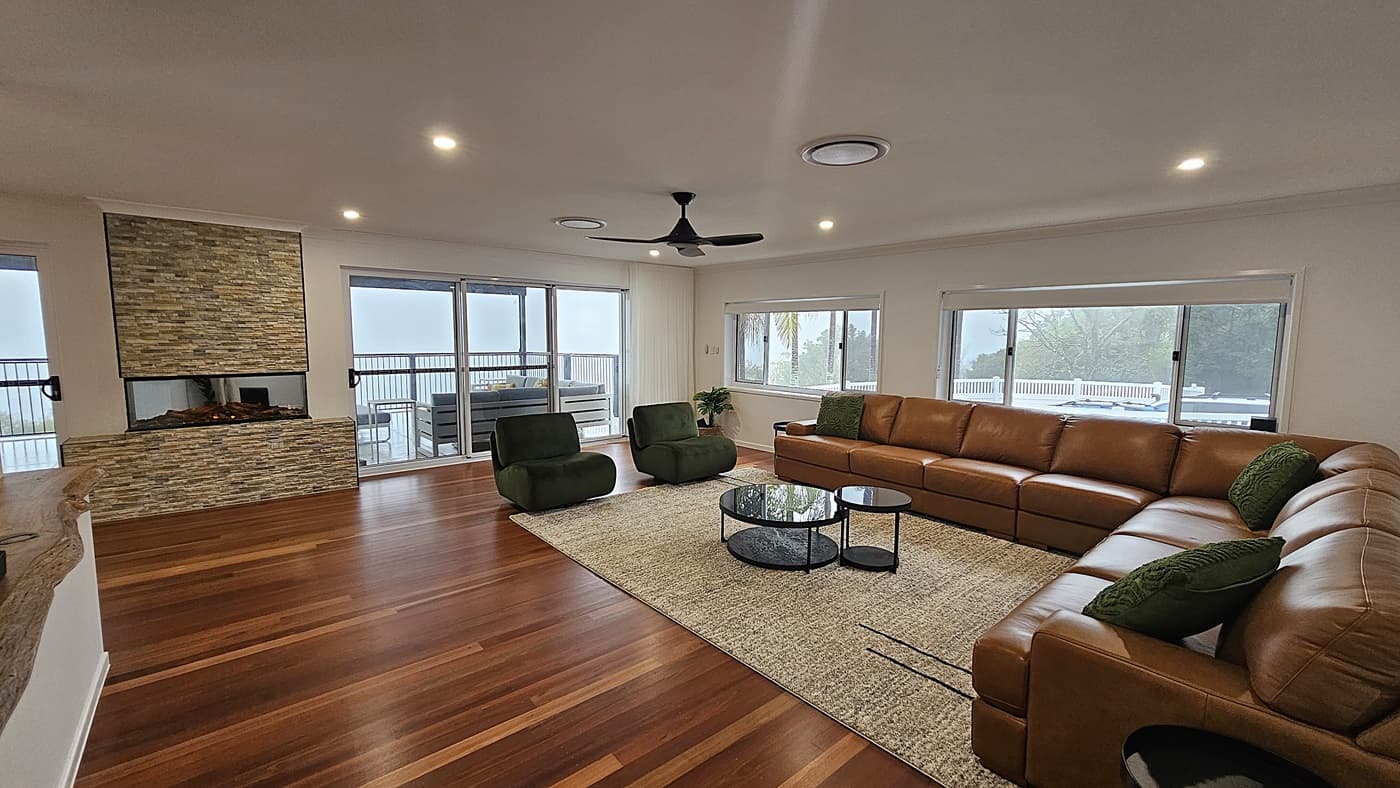 On the left, there is a stone fireplace with a glass front and a sliding glass door leading to an outdoor deck with seating. In the middle, a large tan leather sectional sofa with green cushions sits on a beige rug, accompanied by two green armchairs and a set of round black coffee tables. On the right, large windows with views of trees. The floor is polished wooden planks, and the ceiling is white with recessed lighting, circular air vents, and a black ceiling fan.