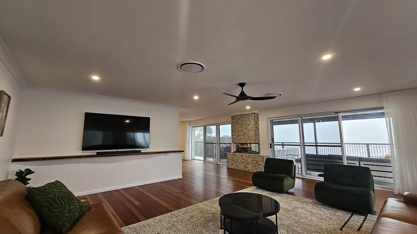 On the left, a large flat-screen TV is mounted on a white wall above a wooden shelf, with a tan sofa and green cushion below. In the middle, a stone fireplace with a glass front stands near sliding glass doors leading to an outdoor deck with seating. On the right, two green armchairs and a round black coffee table sit on a beige rug. The floor is polished wooden planks, and the ceiling is white with recessed lighting, circular air vents, and a black ceiling fan.