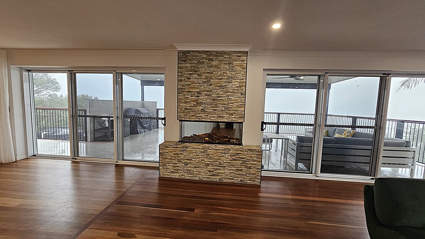 On the left, there are three large sliding glass doors with white curtains, leading to an outdoor deck with a covered barbecue area. In the middle, a stone fireplace with a glass front extends from the floor to the ceiling. On the right, another set of sliding glass doors opens to the same deck, featuring outdoor seating with grey cushions and yellow pillows. The floor is polished wooden planks, and the ceiling is white with recessed lighting.