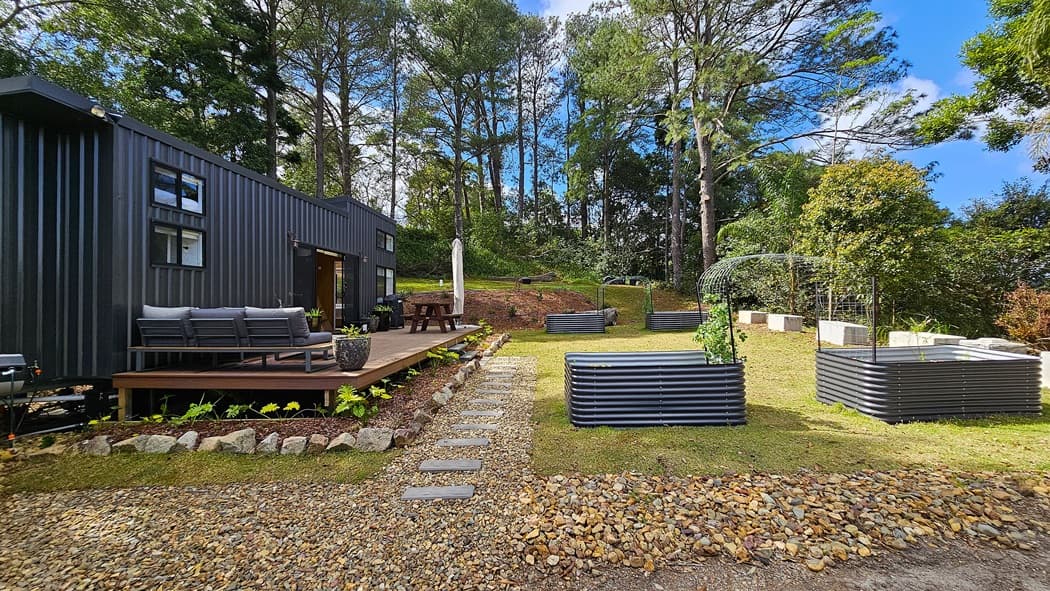 On the left, a black metal building with large windows and a wooden deck furnished with grey outdoor seating. In the middle, a gravel pathway with stepping stones leads to a picnic table with an umbrella. On the right, several raised garden beds made of corrugated metal are arranged on a grassy lawn. The background features tall trees and dense foliage. The floor is a mix of gravel, stepping stones, and grass.