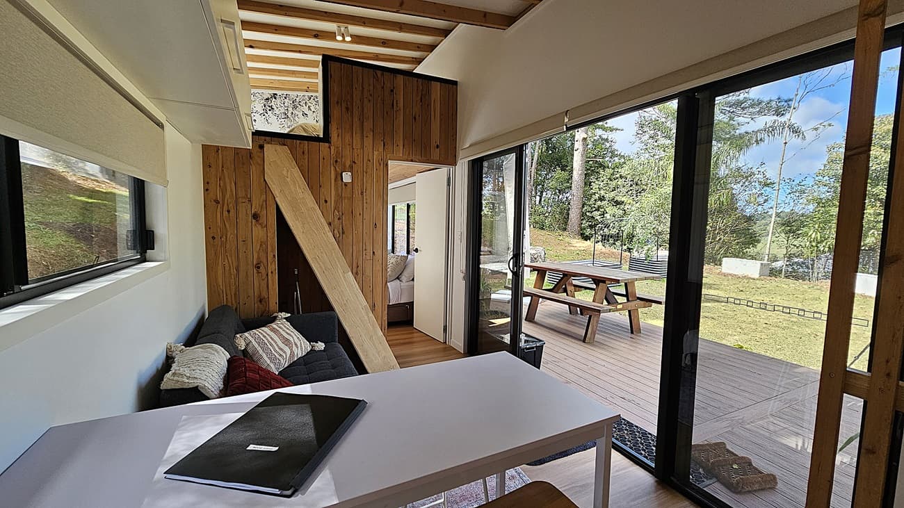 On the left, a white wall with a horizontal window and a grey sofa with cushions. In the middle, a wooden wall with a ladder leading to a loft area, and a doorway to a bedroom. On the right, large sliding glass doors open to a wooden deck with a picnic table. The background features a grassy lawn and tall trees. The floor inside is wooden, and the ceiling has exposed wooden beams.
