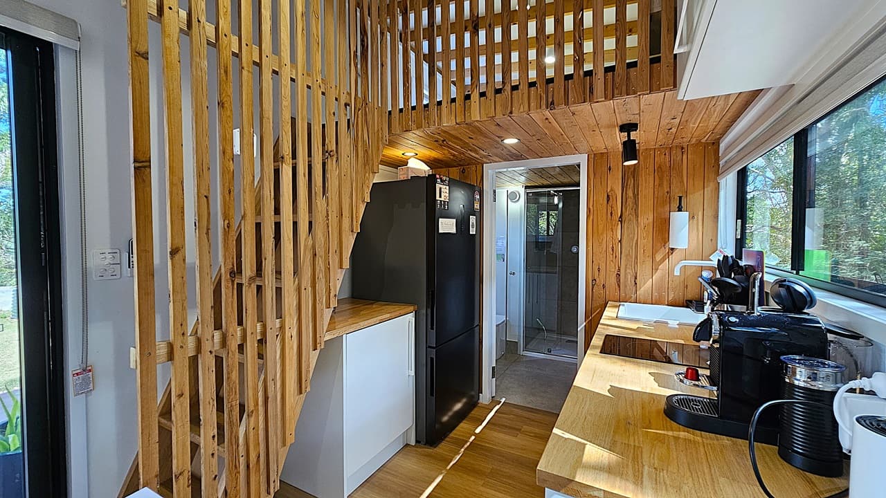 On the left, a wooden staircase with vertical slats leads to an upper loft area. In the middle, a black refrigerator stands next to a doorway leading to a small bathroom with a glass shower. On the right, a wooden countertop with a sink, coffee machine, and kitchen utensils sits below a large window. The background features wooden walls and a view of trees through the window. The floor is wooden, and the ceiling is also wooden with recessed lighting.