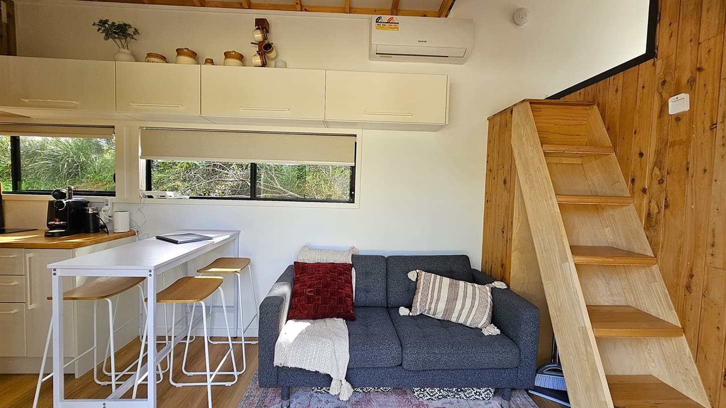 On the left, a wooden countertop with a coffee machine and a white bar table with three wooden stools. In the middle, a grey sofa with cushions and a blanket, positioned under a window with a roller blind. On the right, a wooden staircase leads to a loft area. The background features white walls with upper cabinets and an air conditioner. The floor is wooden, and the ceiling has exposed wooden beams.