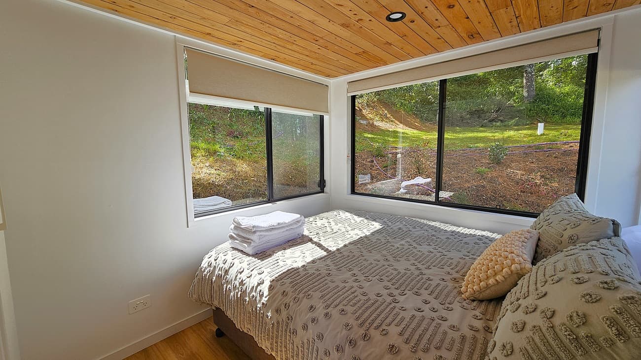 On the left, a white wall with a small power outlet near the floor. In the middle, a bed with a textured beige duvet and several pillows, topped with a stack of folded white towels. On the right, large windows with beige roller blinds offer a view of a grassy slope and trees outside. The background features a green lawn and foliage. The floor is wooden, and the ceiling is made of wooden planks with recessed lighting.