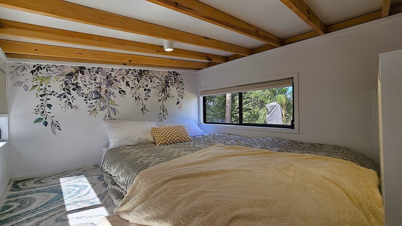 On the left, a white wall with a floral mural featuring green and purple leaves. In the middle, a large bed with a grey patterned duvet, white pillows, and a beige throw blanket. On the right, a rectangular window with a beige roller blind, offering a view of trees outside. The floor is covered with a grey and blue patterned rug. The ceiling has exposed wooden beams and a white surface with a small recessed light.