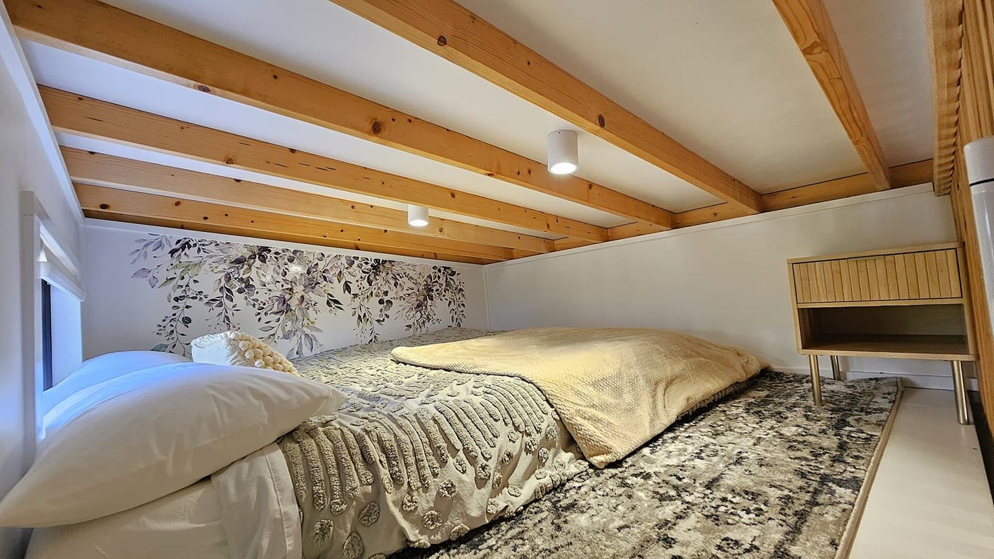 On the left, a white wall with a small window covered by a white roller blind. In the middle, a large bed with a textured beige duvet, white pillows, and a decorative cushion, set against a floral mural with green and purple leaves. On the right, a wooden bedside table with an open shelf. The floor is covered with a grey and white patterned rug. The ceiling has exposed wooden beams and white panels with two small cylindrical light fixtures.