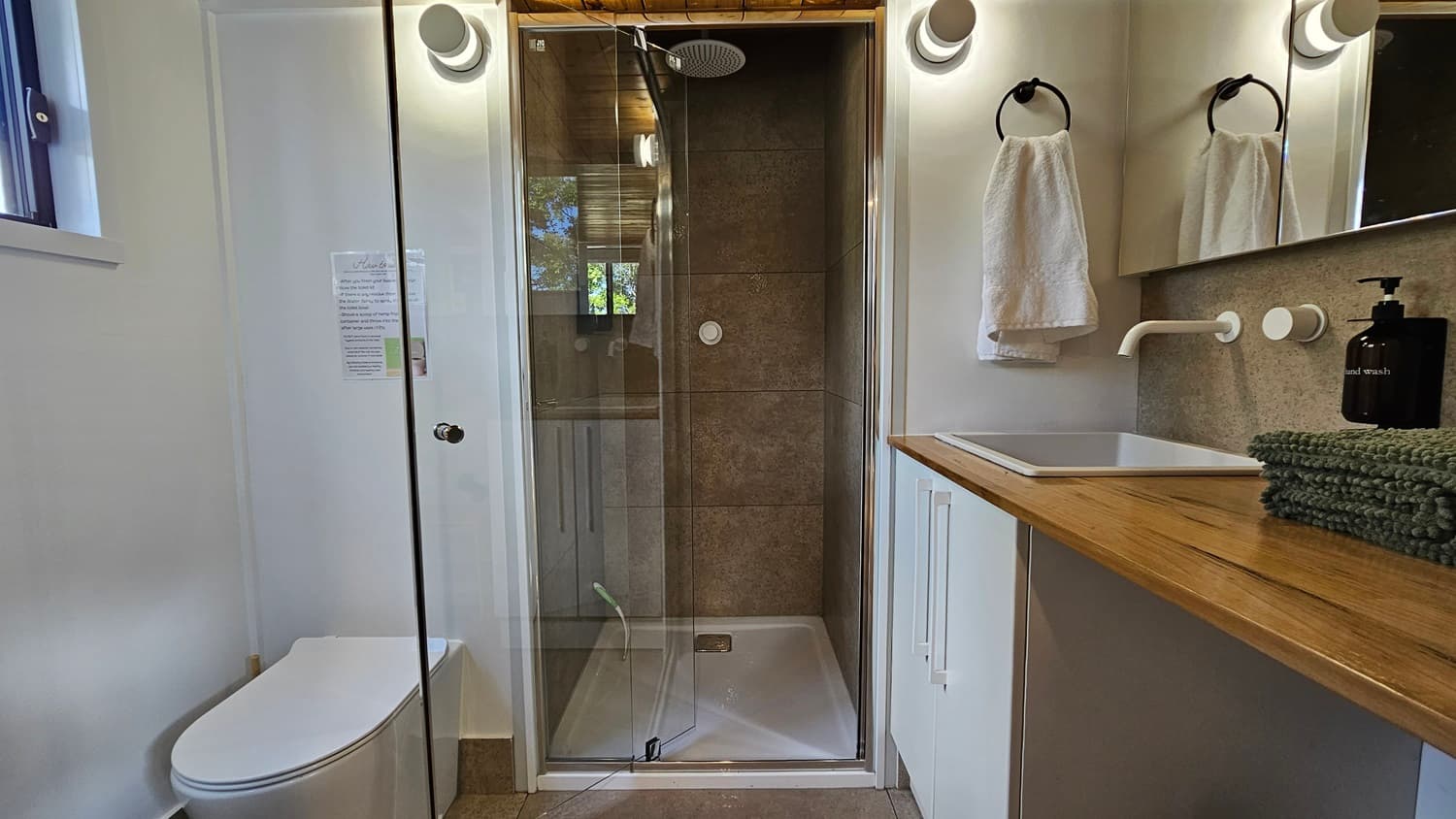 On the left, a white toilet next to a small window with a white frame. In the middle, a glass-enclosed shower with beige tiles and a rain showerhead. On the right, a wooden countertop with a white sink, a black soap dispenser, and a stack of green towels. Above the sink, a mirror with a white towel hanging on a black ring. The floor is tiled, and the ceiling has wooden panels with recessed lighting.