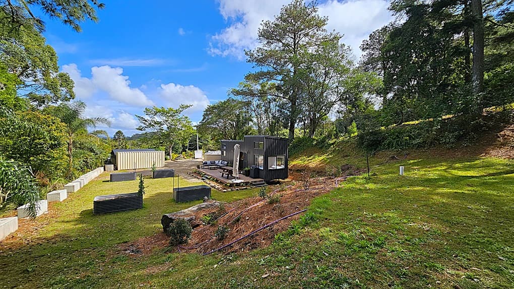 On the left, a row of raised garden beds with small plants, bordered by a low concrete wall. In the middle, a black modern cabin with large windows and a wooden deck furnished with outdoor seating and an umbrella. On the right, a grassy slope with scattered trees and shrubs. The background features dense trees and foliage, creating a forested area. The foreground includes a grassy lawn with patches of dirt and a few small plants.
