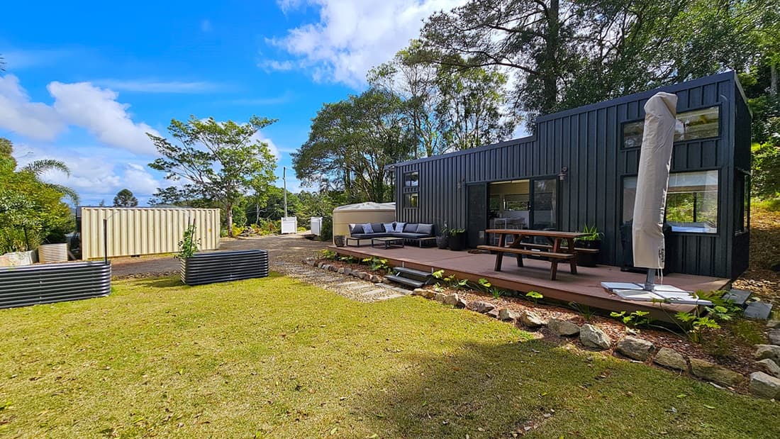 On the left, a beige shipping container and two raised garden beds with small plants. In the middle, a black modern cabin with large windows, a wooden deck, outdoor seating, and a picnic table. On the right, a large umbrella wrapped in a cover stands next to the deck. The background features tall trees and dense foliage. The foreground includes a grassy lawn with a stone border along the deck.
