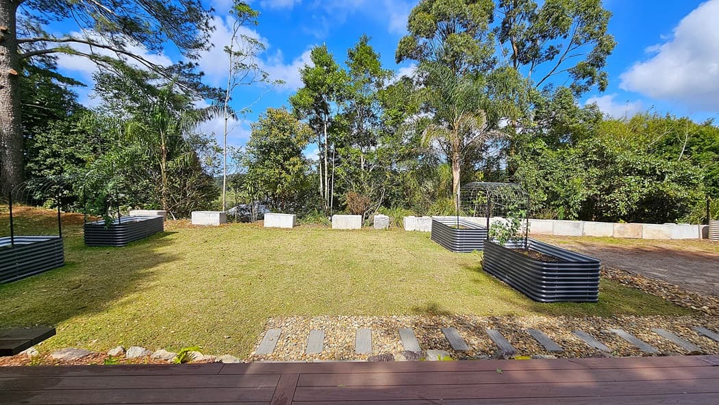 On the left, a raised garden bed with metal sides and small plants. In the middle, a grassy lawn bordered by a row of large white concrete blocks. On the right, another raised garden bed similar to the one on the left, with a metal trellis. The background features dense trees and foliage. The foreground includes a wooden deck with a stone pathway leading into the lawn.