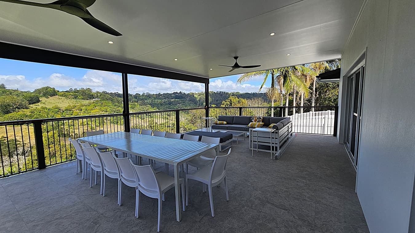 A spacious outdoor patio with a large rectangular dining table and white chairs on the left. On the right, there is a grey sectional sofa with yellow and grey cushions, accompanied by a small coffee table. The background features a scenic view of green hills and trees. The floor is tiled in a light grey colour, and the ceiling is white with recessed lighting and two black ceiling fans. The patio is enclosed with black railings.