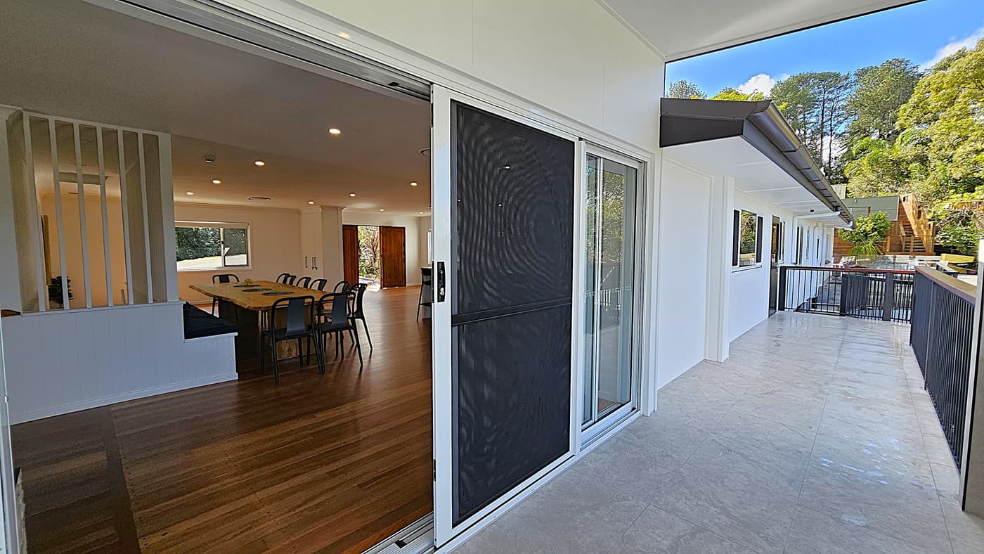 On the left, there is an open-plan interior with wooden flooring, a long dining table surrounded by chairs, and a partition with vertical slats. In the middle, a sliding glass door with a black mesh screen leads to the outside. On the right, there is a spacious tiled balcony with a black railing, white walls, and a view of trees and greenery in the background. The ceiling inside is white with recessed lighting, while the balcony has a white overhang.