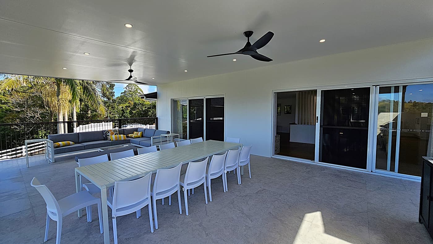 On the left, there is a grey outdoor sofa with yellow and grey cushions, positioned against a black railing with a view of palm trees and greenery. In the middle, a long white dining table with white chairs is set on a light-coloured tiled floor. On the right, there are sliding glass doors leading to an interior space. The ceiling is white with recessed lighting and two black ceiling fans.