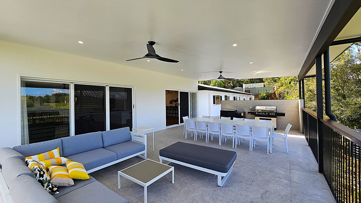 On the left, there is a grey outdoor sofa with yellow and grey cushions, positioned against a white wall with sliding glass doors. In the middle, a long white dining table with white chairs is set on a light-coloured tiled floor. On the right, there is a black railing with a view of trees and greenery. The background features an outdoor kitchen with a barbecue grill. The ceiling is white with recessed lighting and two black ceiling fans.