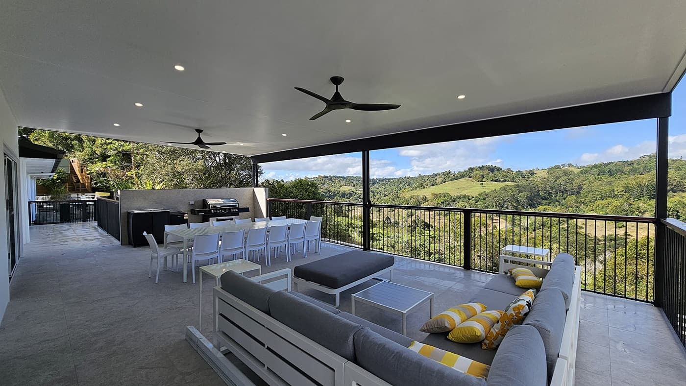 On the left, there is a white wall with sliding glass doors and a black railing. In the middle, a long white dining table with white chairs is set on a light-coloured tiled floor, with a barbecue grill and counter behind it. On the right, a grey outdoor sofa with yellow and grey cushions faces a black railing overlooking a green landscape. The ceiling is white with recessed lighting and two black ceiling fans.