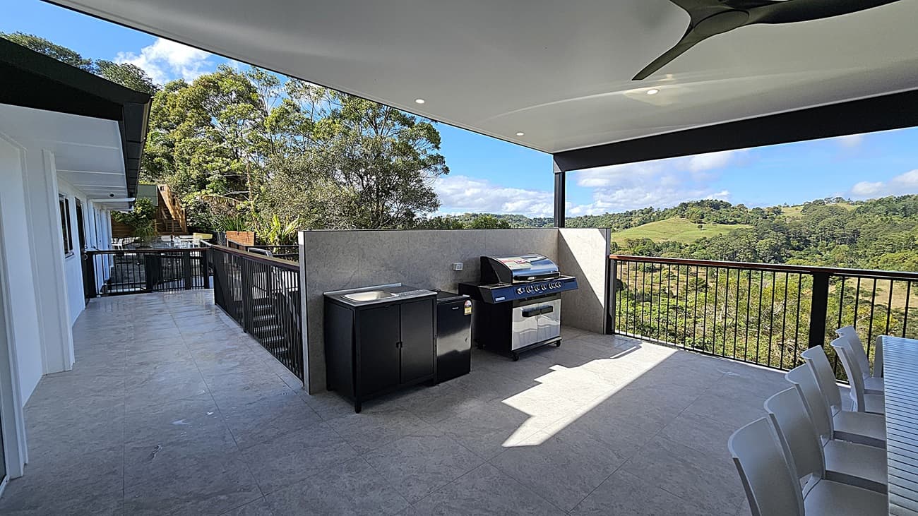 On the left, there is a white wall with windows and a black railing extending along a tiled balcony. In the middle, there is an outdoor kitchen area with a barbecue grill, a sink, and storage cabinets against a grey wall. On the right, a black railing overlooks a green landscape with trees and hills. The foreground features a tiled floor, and the ceiling is white with recessed lighting and a black ceiling fan.