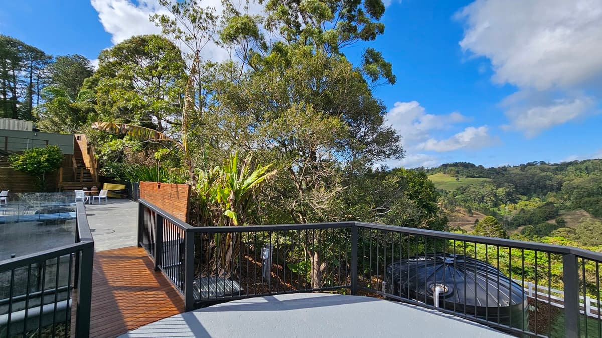 On the left, there is a wooden deck with black railings leading to a concrete patio area with white chairs and a yellow bench. In the middle, tall trees with dense foliage dominate the view. On the right, a black water tank is visible behind black railings, overlooking a green, hilly landscape. The floor is a mix of wooden decking and concrete, and there is no visible ceiling.