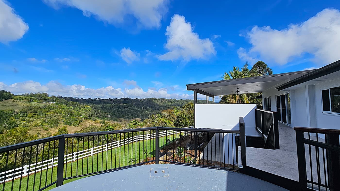 On the left, there is a black railing overlooking a green landscape with trees and hills. In the middle, a white fence runs along the edge of a grassy area. On the right, a white building with large windows and a covered patio area with a ceiling fan is visible. The floor is a mix of grey concrete and light-coloured tiles. The ceiling of the patio is white with a black ceiling fan.