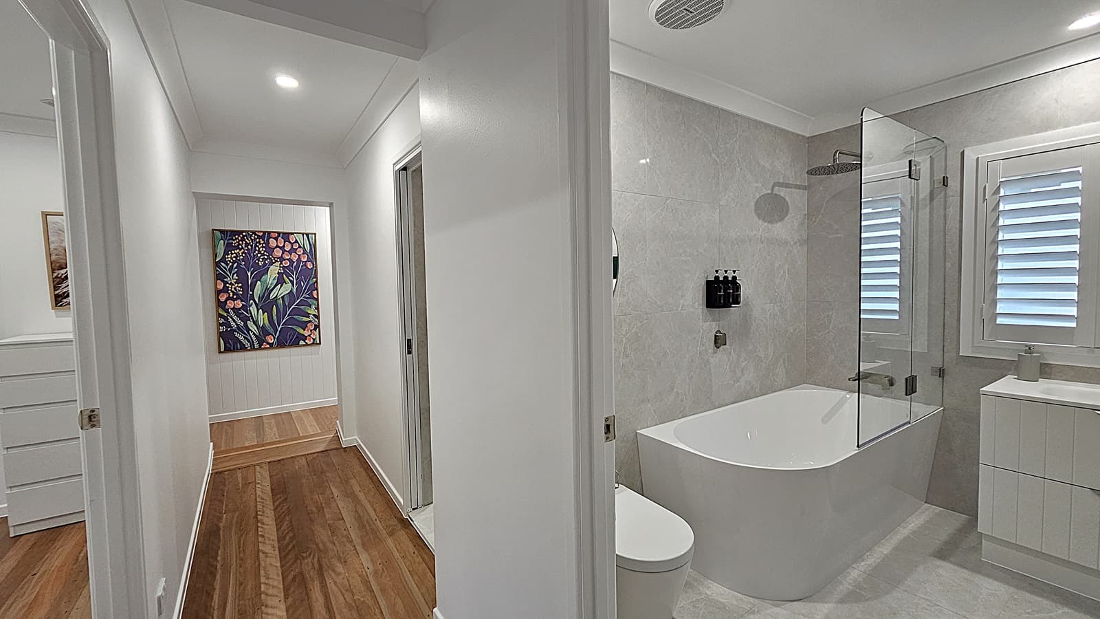 On the left, a hallway with wooden flooring leads to a white chest of drawers and a colourful floral painting on the wall. On the right, a bathroom with grey tiled walls features a white bathtub with a glass shower screen, a wall-mounted showerhead, and a white vanity unit with a sink. The floor in the bathroom is grey tile, and the ceiling is white with recessed lighting and an air vent.