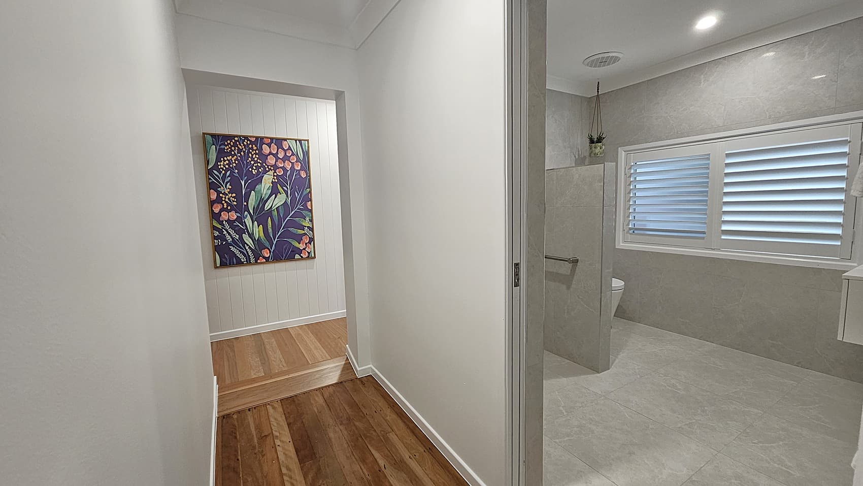 On the left, a hallway with wooden flooring leads to a white panelled wall with a colourful floral painting. On the right, a bathroom with grey tiled walls features a white toilet, a glass shower partition, and a window with white plantation shutters. The floor in the bathroom is grey tile, and the ceiling is white with recessed lighting and an air vent.
