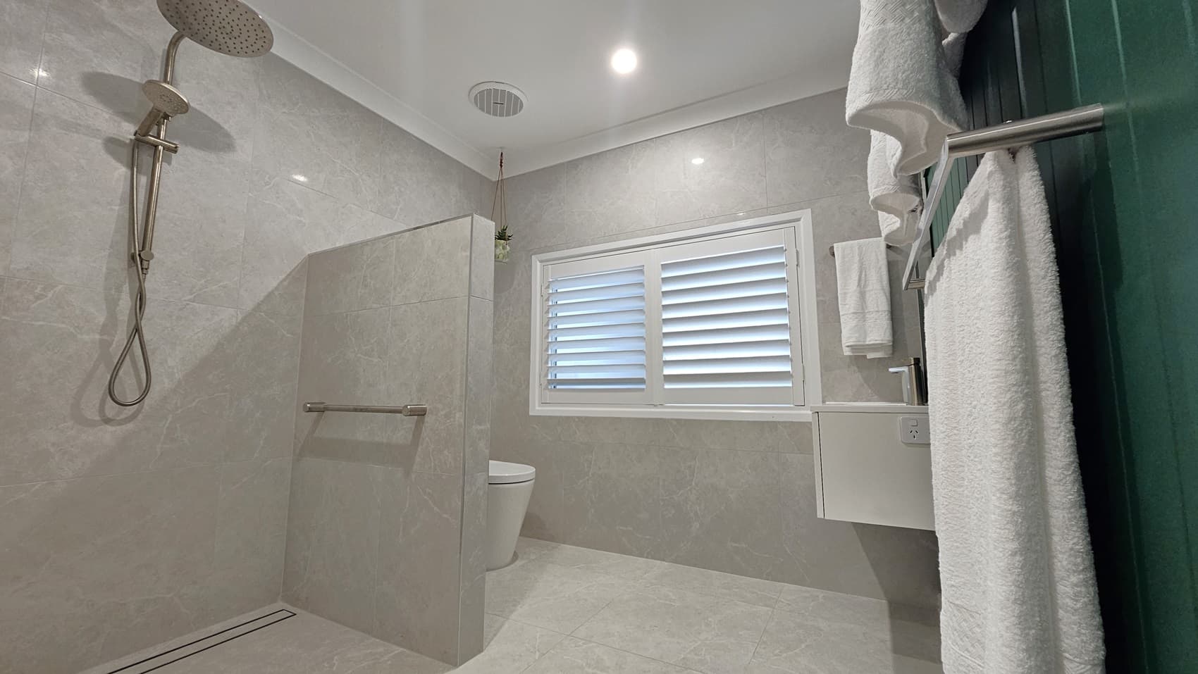 On the left, a wall-mounted showerhead with a handheld attachment above a glass shower partition. In the middle, a white toilet partially visible next to a window with white plantation shutters. On the right, a white vanity unit with a sink, a towel rack with white towels, and a green panelled wall. The floor is grey tile, and the ceiling is white with recessed lighting and an air vent.