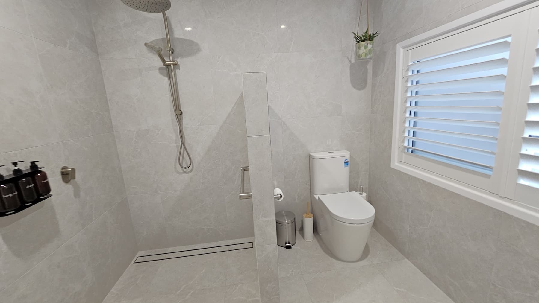 On the left, a wall-mounted showerhead with a handheld attachment and a shelf holding three soap dispensers. In the middle, a partial glass partition separates the shower area from the rest of the bathroom. On the right, a white toilet next to a window with white plantation shutters, and a small hanging plant above. The floor is light grey tile, and the ceiling is white with recessed lighting.