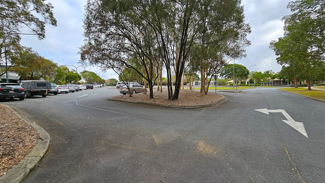 On the left, there is a large, modern building with a grey metal exterior, featuring the words "MORAYFIELD SPORT AND EVENTS CENTRE" prominently displayed. The entrance is flanked by small shrubs and a pathway leading up to it. To the right, a row of trees provides some greenery, with a gravel area and parked cars visible further back. The road is asphalt, smooth and slightly sloped, while the surrounding landscape includes patches of grass and low shrubs.